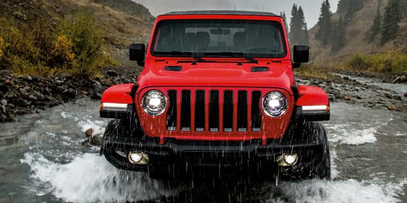 A red Jeep Wrangler driving through a river in a mountainous landscape.