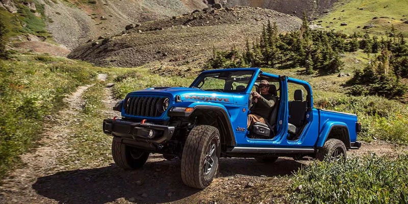 A blue off-road Jeep vehicle driving on a dirt path surrounded by green fields and mountains.