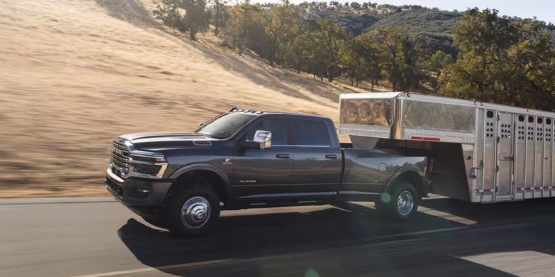 A dark gray RAM pickup truck towing a large trailer on a rural road surrounded by hills and trees.